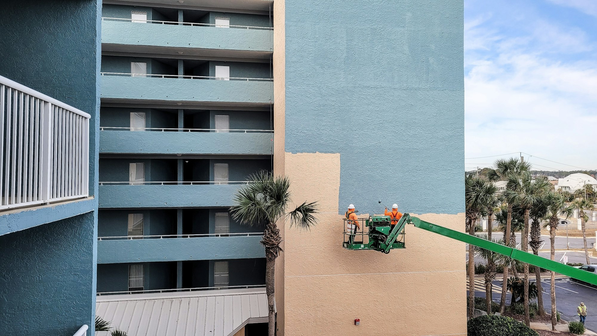 Crane in heavy machinery lifts construction workers up side of building to paint new blue color.